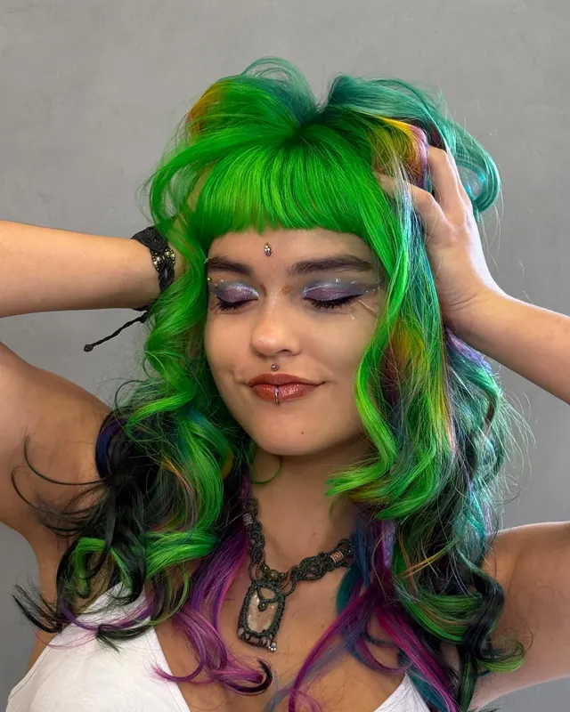 Cabelo longo e ondulado em verde neon com mechas azuis, roxas e rosa, com franja reta, diante de uma parede cinza