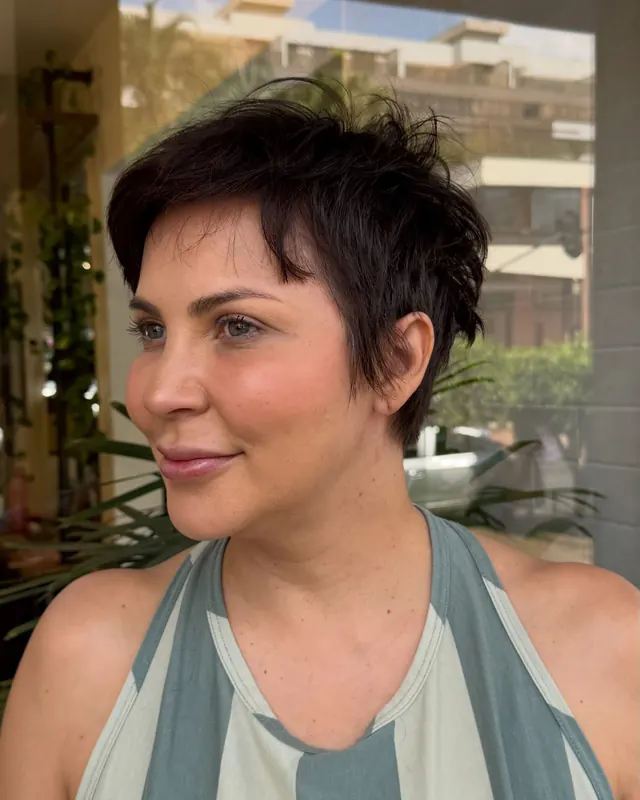Mulher de corte pixie castanho-escuro, com franja desfiada, olhando para o lado, com plantas ao fundo